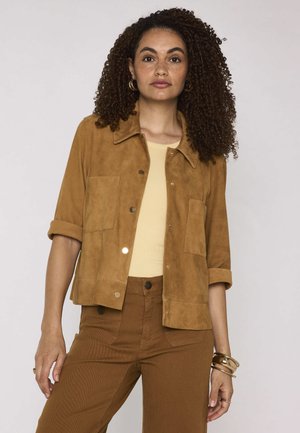 Femme aux cheveux bouclés portant une veste en daim beige sur un haut beige et un pantalon marron taille haute, accessoirisée de boucles d’oreilles créoles dorées et de bracelets.