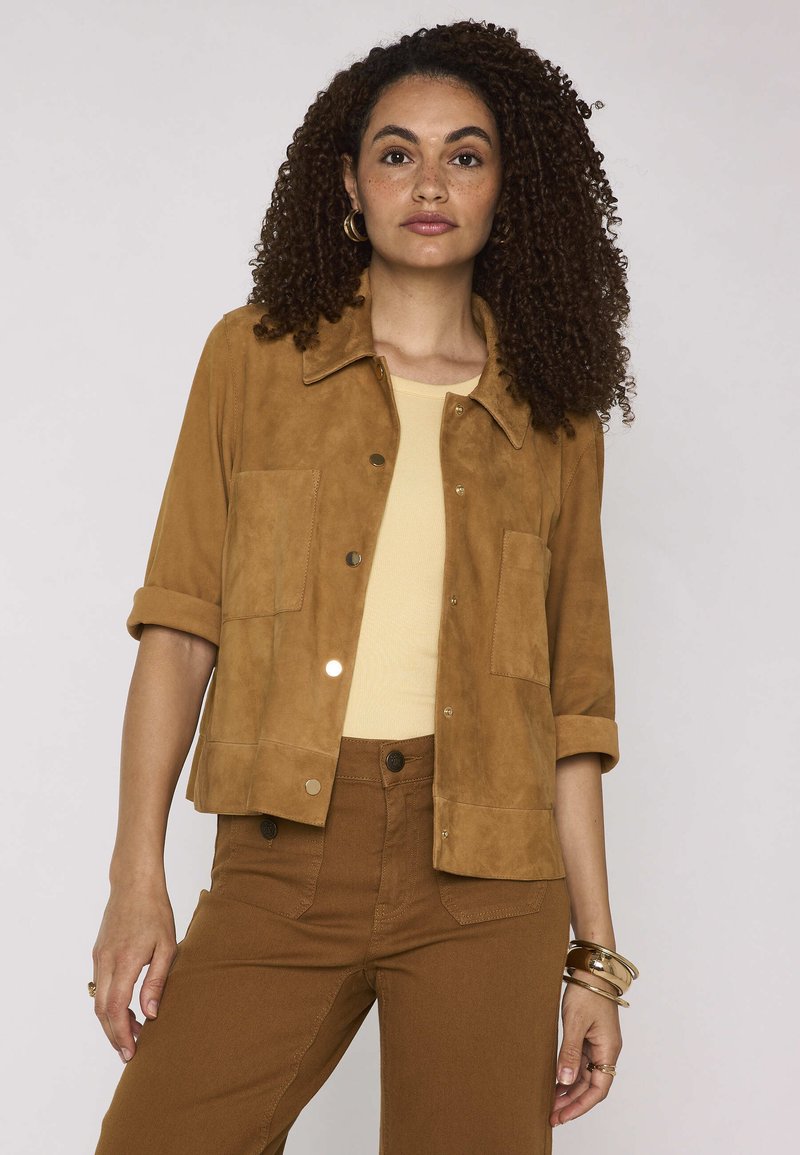 Femme aux cheveux bouclés portant une veste en daim beige sur un haut beige et un pantalon marron taille haute, accessoirisée de boucles d’oreilles créoles dorées et de bracelets.
