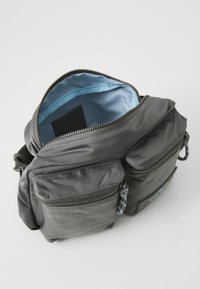 Szara torba z nylonu z zamkami błyskawicznymi, posiadająca liczne zewnętrzne kieszenie oraz niebieską podszewkę z tkaniny. Miękka struktura i funkcjonalny design.
