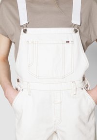 Persoon draagt witte denim tuinbroek met een voorvak en metalen sluitingen over een beige shirt met korte mouwen, handen in de zijkanten van de broekzakken.