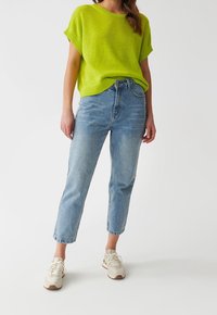 Žena v jasně zeleném krátkém svetru s krátkým rukávem, v světle modrých džínách s vysokým pasem a bílých teniskách, stojící na jednoduchém pozadí.