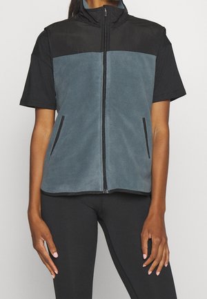 Mujer que lleva un chaleco de forro polar sin mangas con cremallera en gris y negro sobre una camiseta negra de manga corta con mallas negras.