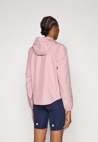 Ανοιχτό ροζ αδιάβροχο μπουφάν με κουκούλα και ελαστικά μανίκια, σε συνδυασμό με σκούρο μπλε σορτς που έχουν δύο λευκές βούλες ως διακοσμητικά στοιχεία.