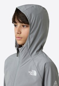 Lys grå hettejakke med glidelås foran, med en hvit logo på brystet og glatt, vannavstøtende stoff.