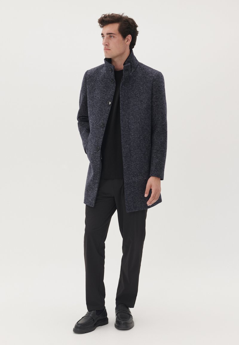 Cappotto in misto lana di un blu navy scuro con colletto alto, chiusura monopetto e due tasche frontali, indossato sopra una camicia nera e pantaloni.