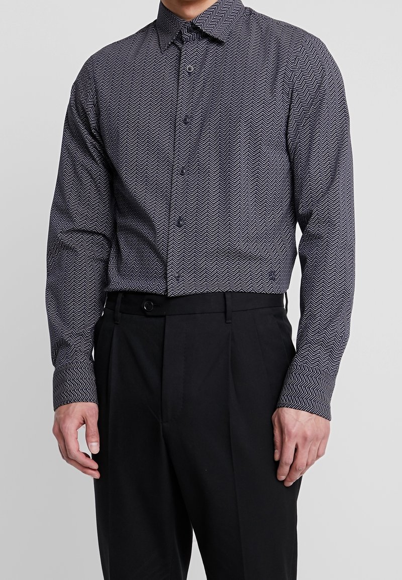 Černobíle vzorovaná košile na knoflíky s herringbonovým designem, se standardním límcem a dlouhými rukávy, doplněná černými kalhotami.