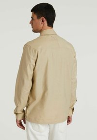 Beige Langarmhemd mit Kragen, das auf der Rückseite eine einzelne Tasche, umknöpfbaren Manschetten und einen strukturierten Stoff aufweist. Weiße Hose sichtbar.