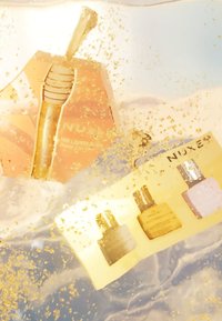 Złote opakowanie z dyszą pompkową i trzema butelkami—złotą, beżową i różową—na miękkim żółtym tle, otoczone migoczącymi cząstkami.