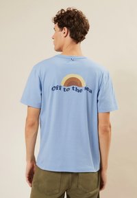 Camiseta de algodón azul claro con cuello redondo y mangas cortas, con una impresión colorida de un arcoíris y texto en la parte trasera.