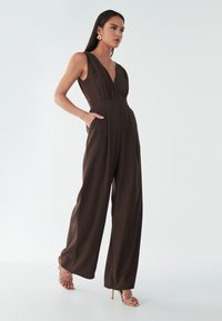 Smeđi kombinezon s dubokim V-izrezom, istaknutim strukom i širokim hlačnim nogavicama. Ima dva bočna džepa i glatku, teksturiranu tkaninu. Zlatne sandale s remenčićima.