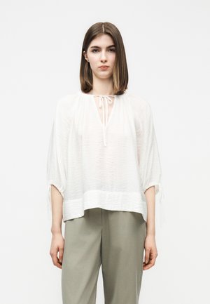 Νεαρή γυναίκα με ίσια καστανά μαλλιά φορά χαλαρό λευκό πουκάμισο με δέσιμο στο λαιμό και ανοιχτό γκρι παντελόνι με φαρδιά μπάρα, στέκεται μπροστά σε απλό φόντο.