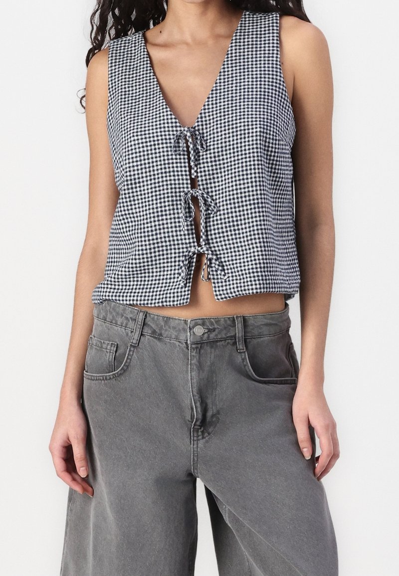 Top corto de cuadritos vichy en blanco y negro con escote en V profundo, cierre frontal con botones y detalle de lazo. Se lleva con pantalones vaqueros grises de talle alto.