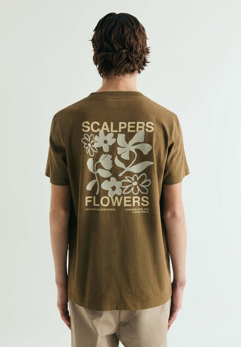 T-shirt en coton vert olive avec un motif floral beige clair au dos, accompagné du texte « SCALPERS FLOWERS » en dessous. Manches courtes et col rond.