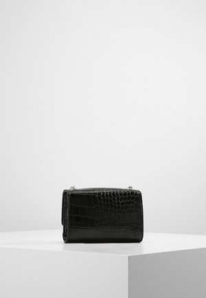 Sac à main en cuir noir à motif crocodile avec des anses en chaîne argentée reposant sur un piédestal géométrique blanc.