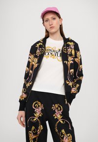 Černá mikina s kapucí na zip s multibarevnou výšivkou květin, bílý tričko s textem "Versace Jeans Couture", ladící černé kalhoty. Růžová kšiltovka.