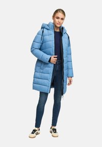 Vrouw staat gekleed in een lichtblauwe gewatteerde pufferjas met capuchon, marineblauwe truien, blauwe skinny jeans en witte sneakers met zwarte strepen.