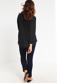 Zwarte blouse met lange mouwen en een losse pasvorm, gecombineerd met donkerblauwe skinny jeans en zwarte ballerina's. Het haar is golvend en valt tot de schouders.