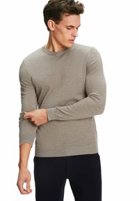 Hellbrauner Strickpullover mit Rundhalsausschnitt, langen Ärmeln und gerippten Bündchen, kombiniert mit dunklen Hosen. Weiche Textur und figurbetontes Design.