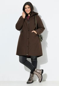 Bruine geïsoleerde parka met capuchon, ritssluiting aan de voorkant, zijzakken en verstelbare trekkoorden, gecombineerd met metallic grijze enkellaarsjes en donkere jeans.