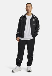 Black athletic tracksuit with white accents, featuring a zip-up jacket and loose-fitting pants. Worn over a white T-shirt, paired with white sneakers.