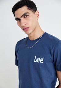 Tricou din bumbac de culoare bleu marin, cu guler rotund și mâneci scurte, având un logo alb "Lee" pe partea stângă a pieptului. Textură fină, croi lejer.