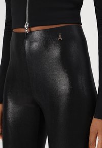 Leggings neri a vita alta realizzati in un materiale lucido e texturizzato, con un piccolo logo vicino alla vita e una zip sulla parte superiore.