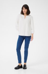 White long-sleeve blouse with a V-neck and lace detailing, paired with blue skinny jeans and black loafers, set against a plain background.