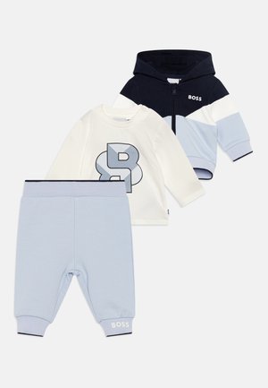 Ensemble de trois articles : hoodie zippé bleu marine, chemise à manches longues blanche avec graphique bleu marine, et pantalon bleu clair avec des poignets côtelés et des détails de logo.