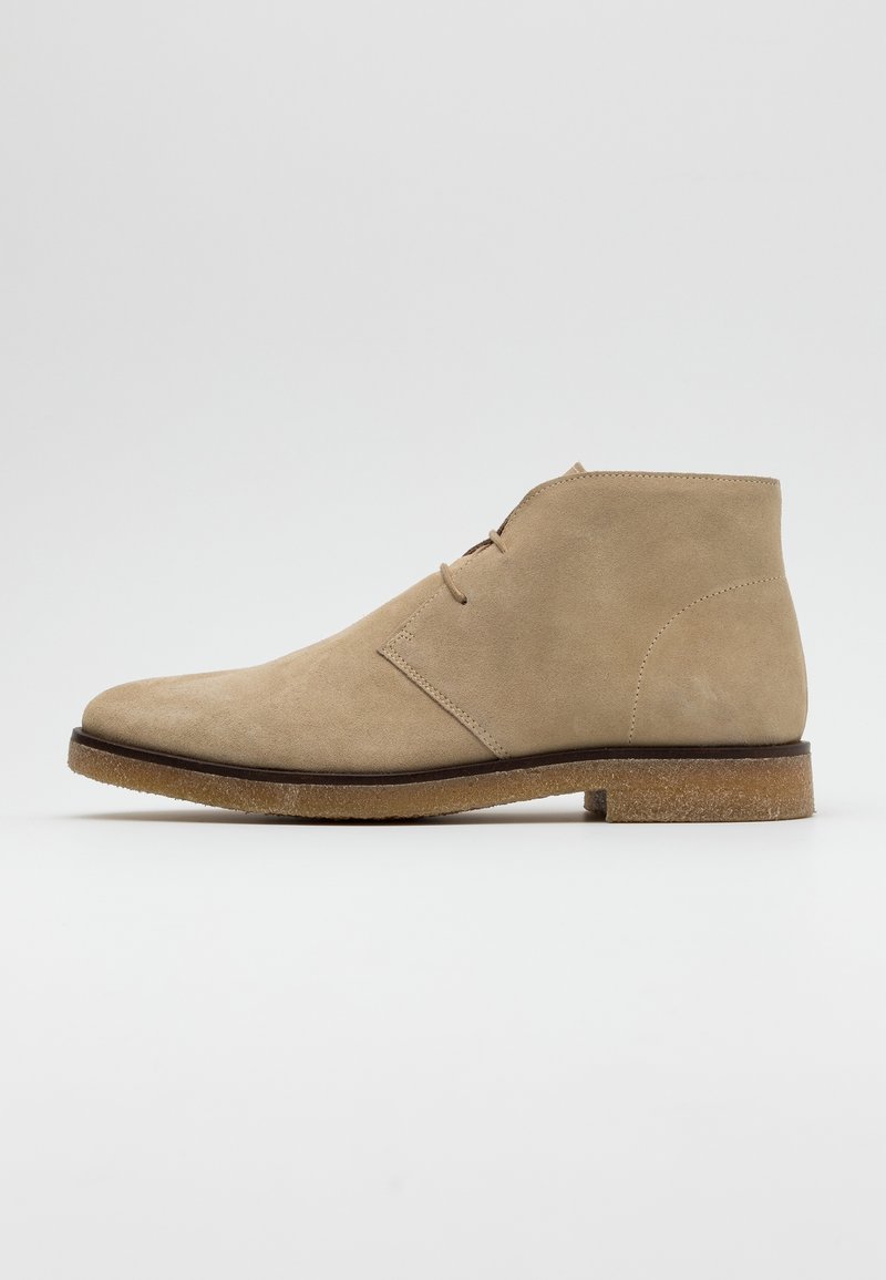 Bottine beige en cuir suédé avec deux œillets pour les lacets, bout arrondi et semelle en crêpe, vue de profil sur fond blanc.