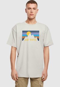 Lyse beige t-shirt med et grafisk motiv af bjerge, en sol og teksten "JUST RHYSE" i gul. Korte ærmer, afslappet pasform, lavet af bomuldsstof.
