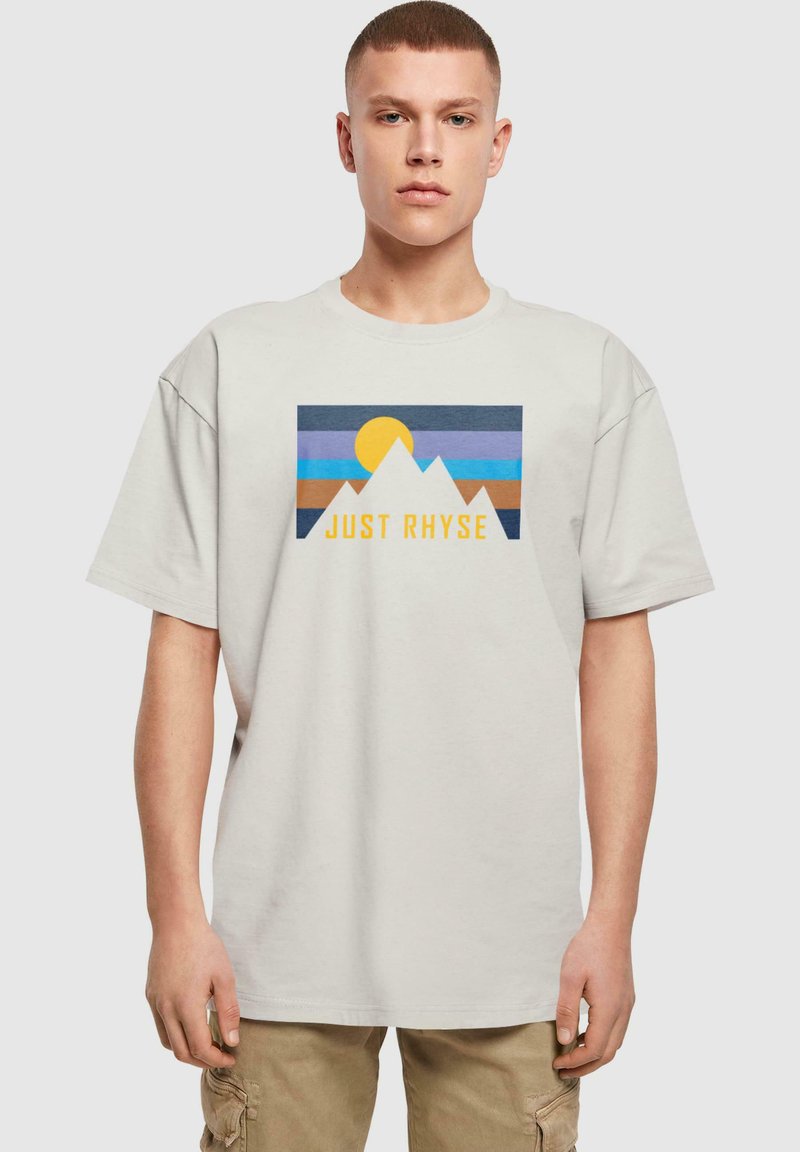 Lyse beige t-shirt med et grafisk motiv af bjerge, en sol og teksten "JUST RHYSE" i gul. Korte ærmer, afslappet pasform, lavet af bomuldsstof.