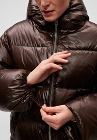 Pruun tepitud jope kõrge kraega, läikiva materjaliga, elastsete mansettidega ja esiosal lukuga. Allääres on reguleerimiseks tõmbenöör.