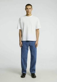 Λευκό κοντομάνικο μπλουζάκι συνδυασμένο με μπλε ίσια τζιν παντελόνια. Το σύνολο περιλαμβάνει μαύρες slip-on παπούτσια και ουδέτερο φόντο.