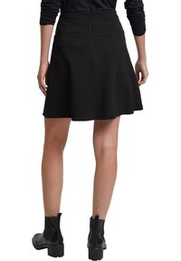 Femme portant une jupe noire évasée aux légères paillettes, un haut noir à manches longues et des bottines noires, vue de dos.