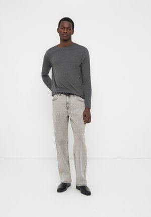 Man standing against white wall wearing dark gray long-sleeve shirt, light gray jeans, black belt, and black shoes.