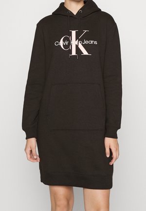 Czarna sukienka z kapturem Calvin Klein Jeans z przednią kieszenią kangurką i różowym logo "CK" na klatce piersiowej, noszona przez osobę od szyi do kolan.
