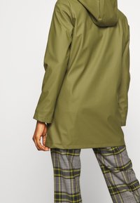 Manteau imperméable vert olive avec capuche, coupe droite et manches longues. Associé à un pantalon à motifs écossais gris et jaune.