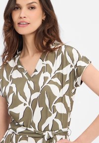 Olivgrünes Kleid mit weißem Blattmuster. Verfügt über einen V-Ausschnitt, kurze Ärmel und einen Gürtel zum Binden in der Taille, gefertigt aus einem glatten Stoff.