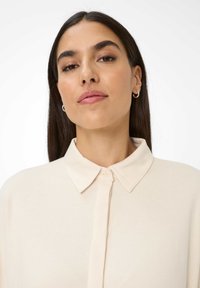 Une femme aux longs cheveux noirs porte de petites boucles d'oreilles en argent et une chemise beige boutonnée avec un col classique, le tout sur un fond blanc.