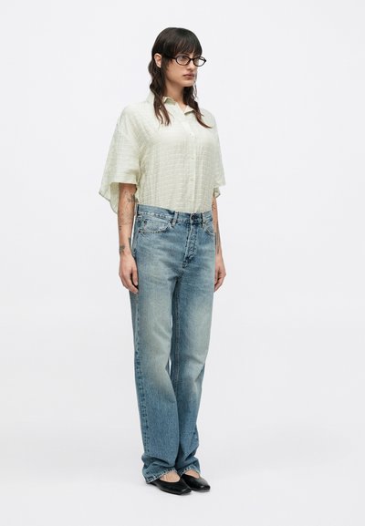 Világoszöld, texturált, gombos ing rövid ujjúval, halványkék, egyenes szabású farmerrel párosítva. A fekete balerina cipők teszik teljessé a szettet.