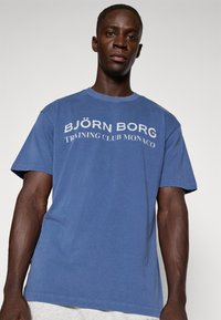 Modré bavlněné tričko s kulatým výstřihem a krátkými rukávy. Na přední straně je text "BJÖRN BORG TRAINING CLUB MONACO" v bílé barvě. Hladký povrch.