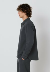 Mladenič s kodrastimi lasmi stoji v profilu, oblečen v temno sivo srajco z dolgimi rokavi in ujemajoče hlače, z rokama v žepih, pred enostavno ozadje.