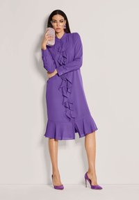 Frau mit langen dunklen Haaren, die ein lila, gerüschtes Kleid und hohe Schuhe trägt, hält eine rosa Glitzer-Clutch nahe ihrem Gesicht und steht vor einer schlichten Wand.