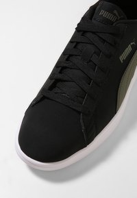 Zapato deportivo negro con parte superior texturizada, lengüeta acolchada y cordones. Presenta un logo verde y un panel lateral, con una suela de goma blanca.
