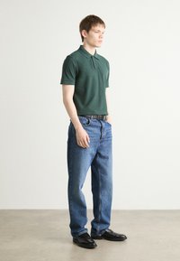 Grønn kortermet polo-skjorte med krage, paret med lyseblå rett-lags jeans og svarte pensko, stående mot en nøytral bakgrunn.