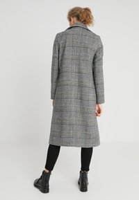 Skräddarsydd lång kappa i grått rutmönster med gula detaljer, gjord av vävd textil. Har krage och sidoinsnitt, kombineras med svarta leggings och ankelstövlar.