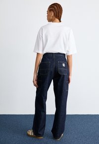 Dark blue denim work pants with a straight-leg design, featuring multiple pockets and yellow stitching. Paired with a white oversized T-shirt.