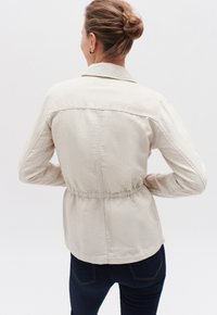 Giacca beige, leggera, in misto cotone con vita arricciata, colletto con bottoni e maniche lunghe. Texture liscia, senza motivi visibili.
