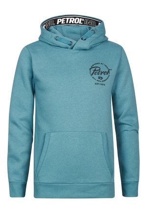 Person iført teal hættetrøje med frontlomme og marineblå jeans, stående mod en ensfarvet hvid baggrund.