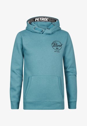 Person iført teal hættetrøje med frontlomme og marineblå jeans, stående mod en ensfarvet hvid baggrund.